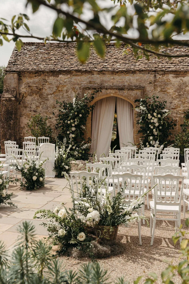 An outdoor ceremony at Caswell House with Cotswold stone, white floral arch and green foliage